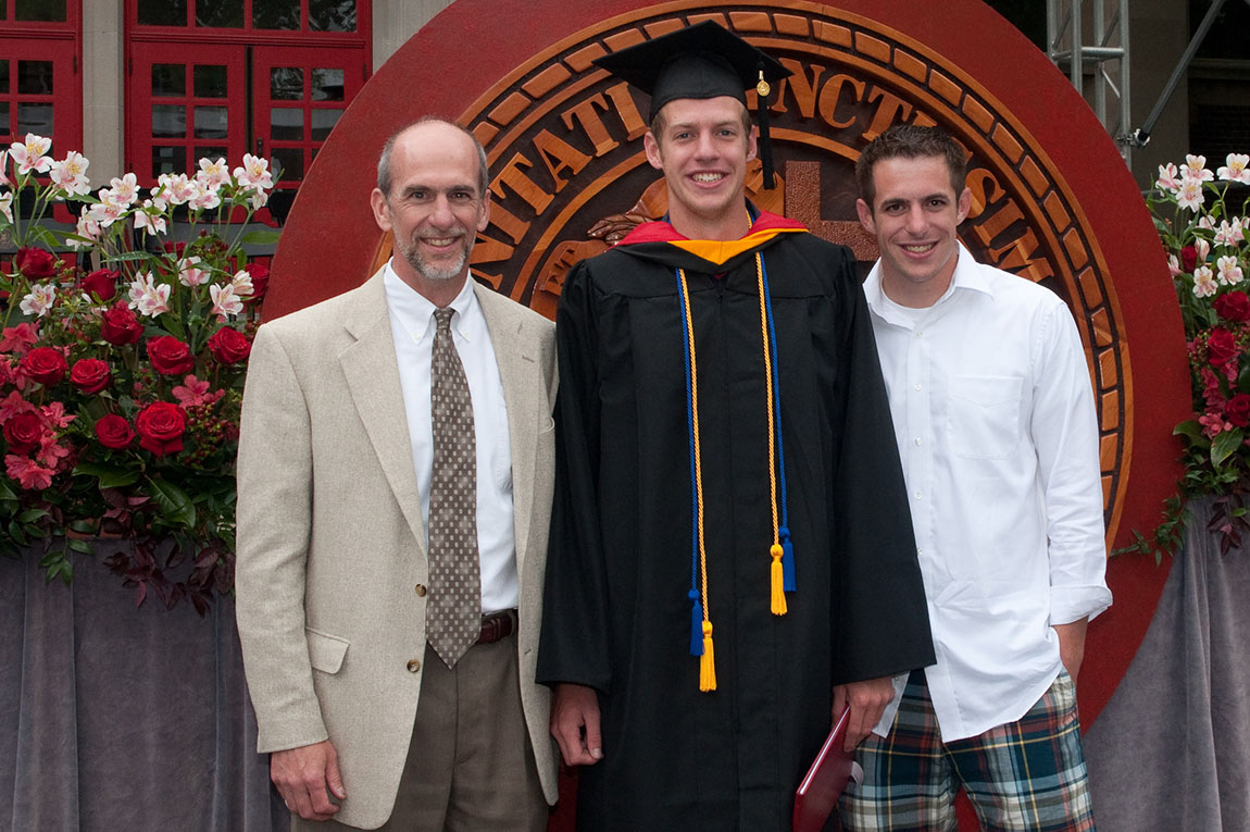 A man in a tan jacket and tie stands next to a college student in a gap and gown and a younger man in a white button down shirt